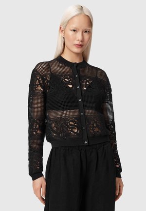 Vrouw met recht, platinumblond haar draagt een zwarte, met knopen gesloten mesh cardigan over een zwarte top en losse zwarte broek, staand tegen een grijze achtergrond.