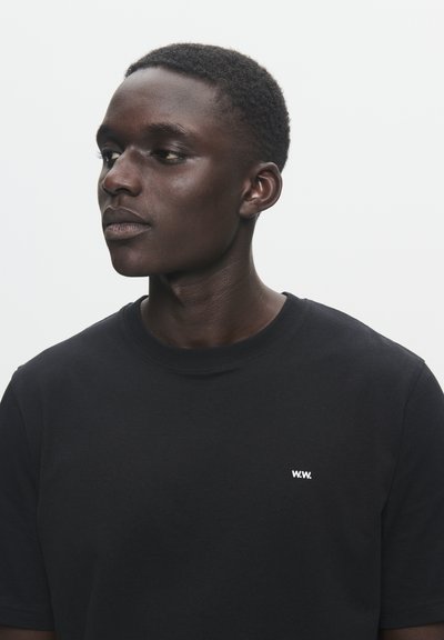 T-shirt de algodão preto com mangas curtas e decote redondo. Apresenta um pequeno logotipo branco na área do peito esquerdo. Textura suave, ajuste descontraído.