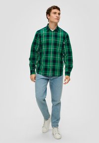 Grün karierte Hemd mit Knöpfen, das einen Kragen, lange Ärmel und weiße Akzente aufweist, kombiniert mit hellblauen Jeans und weißen Sneakers.