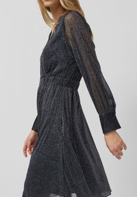 Donkere, sprankelende jurk met een aangesloten taille, lange doorschijnende mouwen, knoopmanchetten en een zwierige rok. Gemaakt van gestructureerde, glanzende stof.