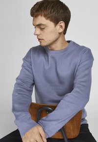 Sudadera lila claro con cuello redondo y mangas largas, exhibida en una persona sentada que descansa sobre una silla de madera. Textura de tela suave.