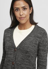 Cardigan en tricot gris avec un col en V, présentant un tissu texturé et deux boutons marron à l'avant, superposé sur une chemise de couleur claire.