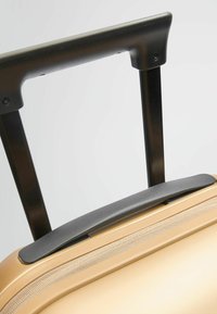 Valise rigide avec une finition dorée métallique, présentant une surface texturée, une poignée noire rétractable et une fermeture éclair sur le bord.