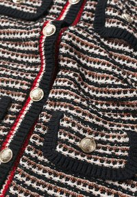 Gestrickte Strickjacke in Schwarz, Weiß und Braun mit horizontalen Streifen, ausgestattet mit goldenen Knöpfen und roten Akzenten entlang der vorderen Öffnung und der Taschen.