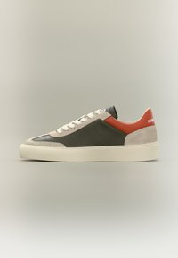 Baskets basses avec semelle blanche, daim beige, panneaux en cuir vert foncé, lacets blancs et languette de talon rouge.