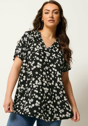 Kvinde iført en sort V-hals bluse med hvide margeritprint og korte ærmer, kombineret med blå jeans mod en ensfarvet baggrund.