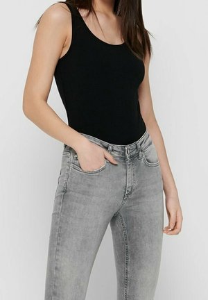 Kobieta ubrana w czarny, bezrękawnik i dopasowane jasnoszare jeansy, z jedną ręką w przedniej kieszeni, na jednolitym tle.