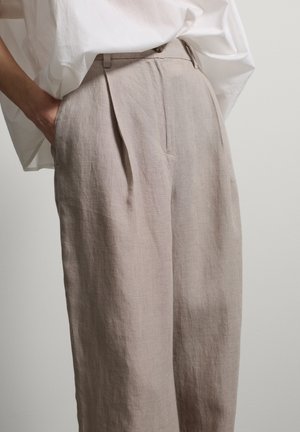 Pantalones de lino beige de talle alto y pierna ancha con bolsillos laterales, usados con camisa blanca por fuera, mano en el bolsillo, sobre un fondo liso.