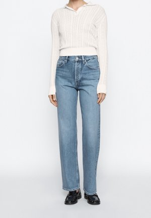 Person iført hvid langærmet striktrøje, lyseblå højtaljede jeans med lige ben og sorte loafers, stående mod en hvid baggrund.