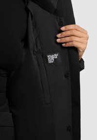 Chaqueta acolchada negra con una textura suave, que presenta una etiqueta de marca y cierre de botones a presión, exhibida con una mano sosteniendo el tejido.