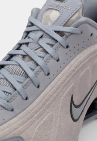 Pilkas Nike Shox sportbačių modelis su tekstūruota zomšos apdaila, tinklelio detalėmis ir ryškiu swoosh logotipu. Šnurrės suderinta pilka spalva su lygia apdaila.
