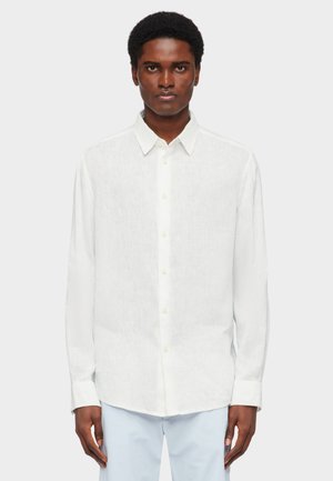 Mann trägt ein weißes Hemd mit langen Ärmeln und hellblaue Hose, steht vor einem schlichten hellgrauen Hintergrund.