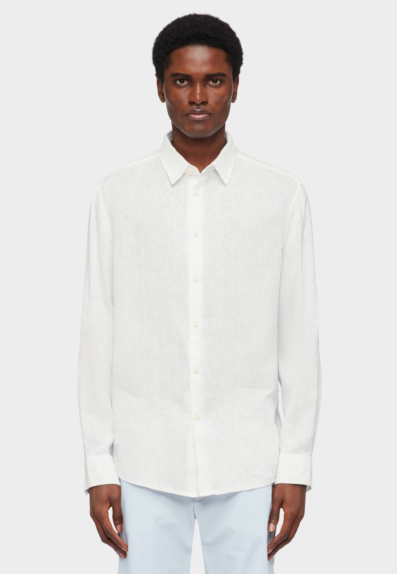 Homme portant une chemise blanche à manches longues boutonnée et un pantalon bleu clair, debout devant un fond gris clair uni.