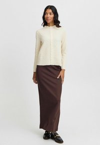 Ljus krämfärgad texturerad långärmad topp med hög krage, tillsammans med en figurnära brun maxikjol och svarta skor med guldkant.