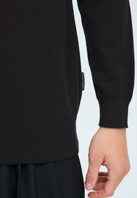 Črn pleteni pulover z rebrastim robom in manšetami. Ima manjšo oznako blagovne znamke na strani. Gladka tekstura, prilegajoča se zasnova.