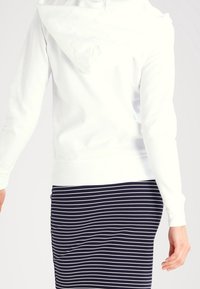Persona con una sudadera blanca y una falda negra con rayas horizontales blancas, vista desde la espalda y a la altura de la cintura.