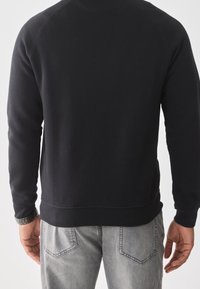 Sudadera negra con puños y dobladillo acanalados, con un diseño sencillo. Combinada con vaqueros gris claro que muestran detalles en los bolsillos traseros.