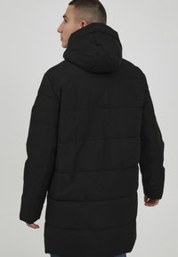 Sorte puffjakke med hette, med et quiltet design. Stoffet ser glatt ut med en matt finish, som strekker seg til midten av låret.