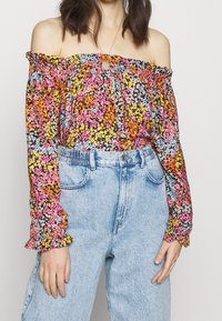 Off-the-shoulder top met een multicolore bloemenpatroon op een zwarte achtergrond; gecombineerd met lichtblauwe high-waisted denim jeans.