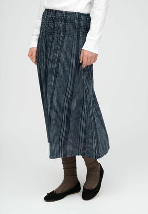 Personne portant une jupe plissée à motif noir et blanc, des chaussettes brunes côtelées, des ballerines noires et un haut blanc à manches longues, debout.