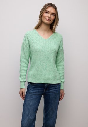 Kvinna med blont hår som bär en ljusgrön stickad tröja och blå jeans, stående mot en enkel ljusgrå bakgrund.