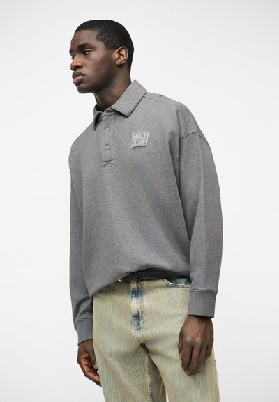 Άνδρας με γκρι πόλο μπλούζα μακρυμάνικη, με λογότυπο στο στήθος και τζιν ανοιχτού χρώματος, στέκεται μπροστά σε λευκό απλό φόντο.