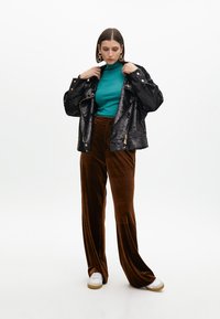 Giacca nera con paillettes sopra un dolcevita teal, abbinata a pantaloni a gamba larga in velluto marrone e sneakers bianche. Orecchini a cerchio dorati completano il look.