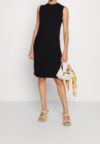 Schwarzes ärmelloses Kleid mit vertikaler gerippter Textur, knielang. Kombiniert mit einer weißen Handtasche und gelben Sandalen, akzentuiert durch einen bunten Schal.