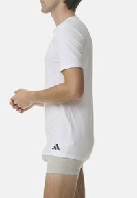 T-shirt bianco a maniche corte in cotone; presenta un logo Adidas nero sul lato sinistro in basso; indossato con boxer grigi.