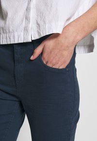 Pantalones azul marino con una textura suave, que presentan bolsillos laterales y un acento de botón visible, combinados con una blusa blanca clara y texturizada.