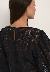 Sort bluse i blonde med blomsterprint, puffærmer og en nøglehulslukning med knapper bagpå. Struktureret stof med et fint udseende.