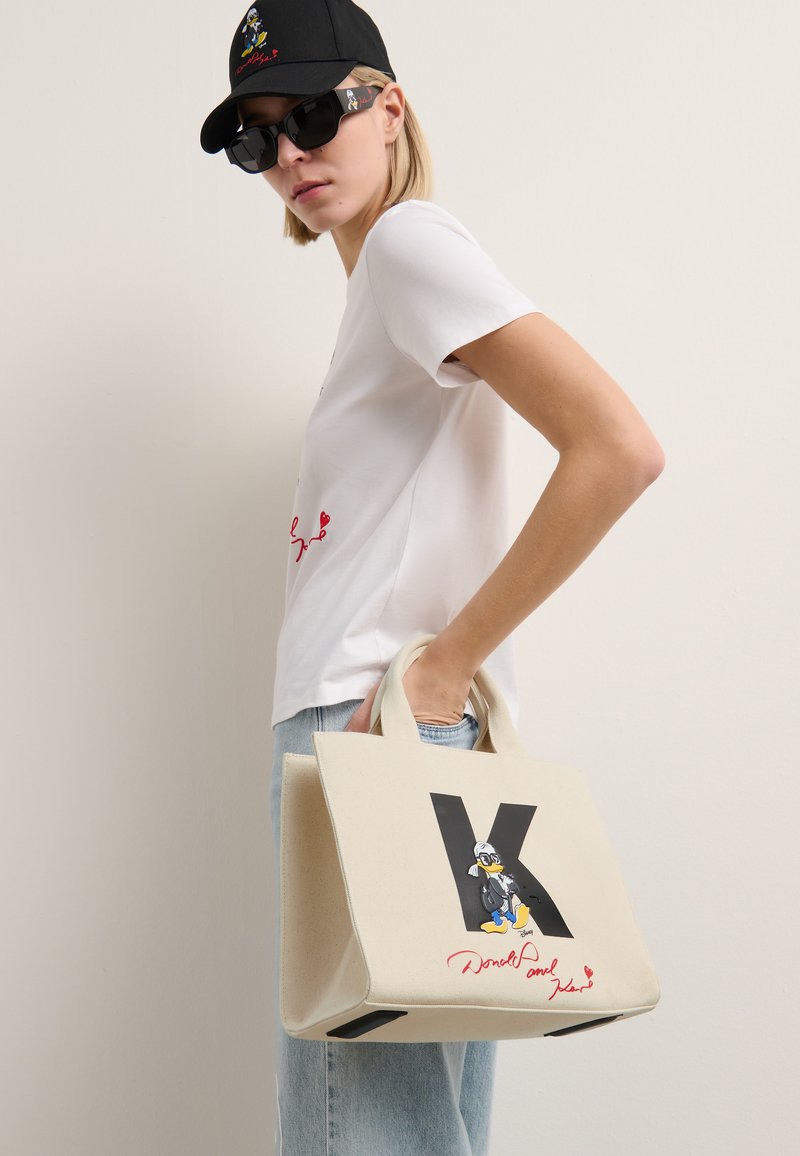 Sac en toile de couleur crème avec un grand "K" noir, mettant en avant un graphique de Donald Duck et du texte rouge. Forme structurée; courtes poignées; design minimaliste.