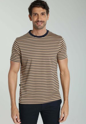 Homme aux cheveux courts et foncés portant une chemise à manches courtes beige et noire rayée et un pantalon noir, debout devant un fond clair uni.