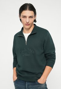 Tumši zaļš džemperis ar rāvējslēdzēju ap kaklu, ar iepresētiem tekstiem "Originals Studio", ribotām piedurknēm un brīvu piegriezumu. Nēsāts virs baltas krekla.