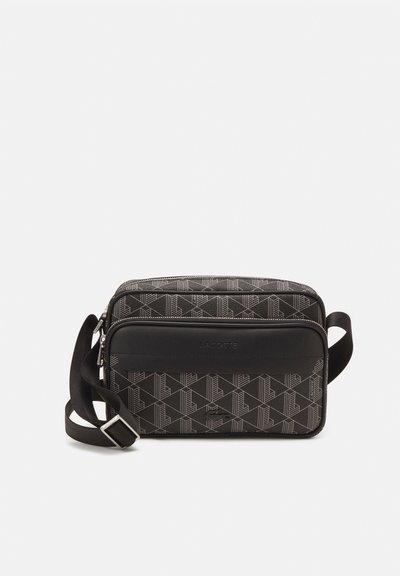 Sac bandoulière noir à motif géométrique avec un panneau en cuir noir lisse, fermeture éclair et sangle réglable. Présente le logo de la marque sur le devant.