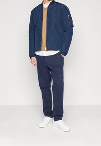 Tmavě modrá prošívaná bunda s kapsou na zip, pod ní béžový svetr, bílá košile, tmavě modré kalhoty a bílé tenisky.