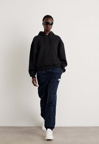 Zwarte oversized hoodie, marineblauwe sportbroek met logo, witte sneakers en donkere zonnebril gedragen door een persoon die tegen een neutrale achtergrond loopt.
