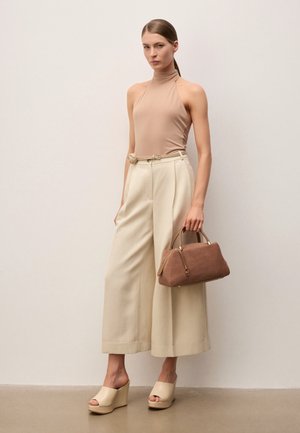 Femme portant un haut beige sans manches, un pantalon ample crème, des sandales compensées beiges, tenant un sac à main en cuir marron devant un fond uni.