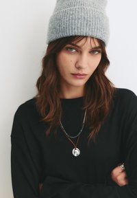 Beanie a coste grigio, maglietta nera a maniche lunghe, collane sovrapposte con perline e un pendente circolare. Capelli lisci, sfondo neutro.