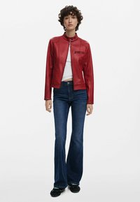 Rote Lederjacke mit hohem Kragen,Reißverschluss vorne und kleiner Brusttasche, kombiniert mit ausgestellten dunkelblauen Jeans und schwarzen Schuhen.