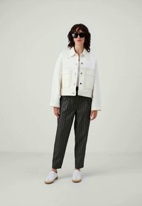 Donna che indossa occhiali da sole neri, giacca di jeans bianca, pantaloni a righe nere e sneakers bianche, in piedi di fronte a uno sfondo neutro.