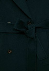 Mörkgrön ullkappa med en dubbelknäppt design, som har ett bälte i midjan, svarta knappar och textil med struktur.