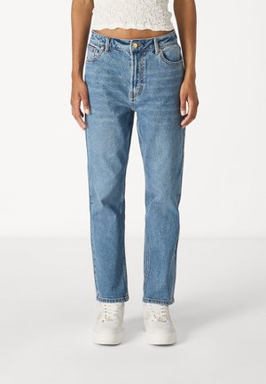 Vrouw draagt lichtblauwe high-waist rechte jeans, witte sneakers en een cropped witte textuurtop, staand tegen een effen achtergrond.