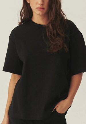 Ung kvinde med langt brunt hår iført en simpel sort oversized T-shirt, den ene hånd i lommen, neutral baggrund.