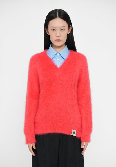 Pull en V, fluffy et rose, fabriqué en matériau doux, avec des manches longues et un accent de logo sur l'ourlet, associé à une chemise bleue à col.