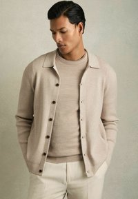 Beige cardigan med ribbat mönster, knappar och krage, lagerpålagd över en matchande polotröja. Ljust färgade byxor fulländar outfiten.