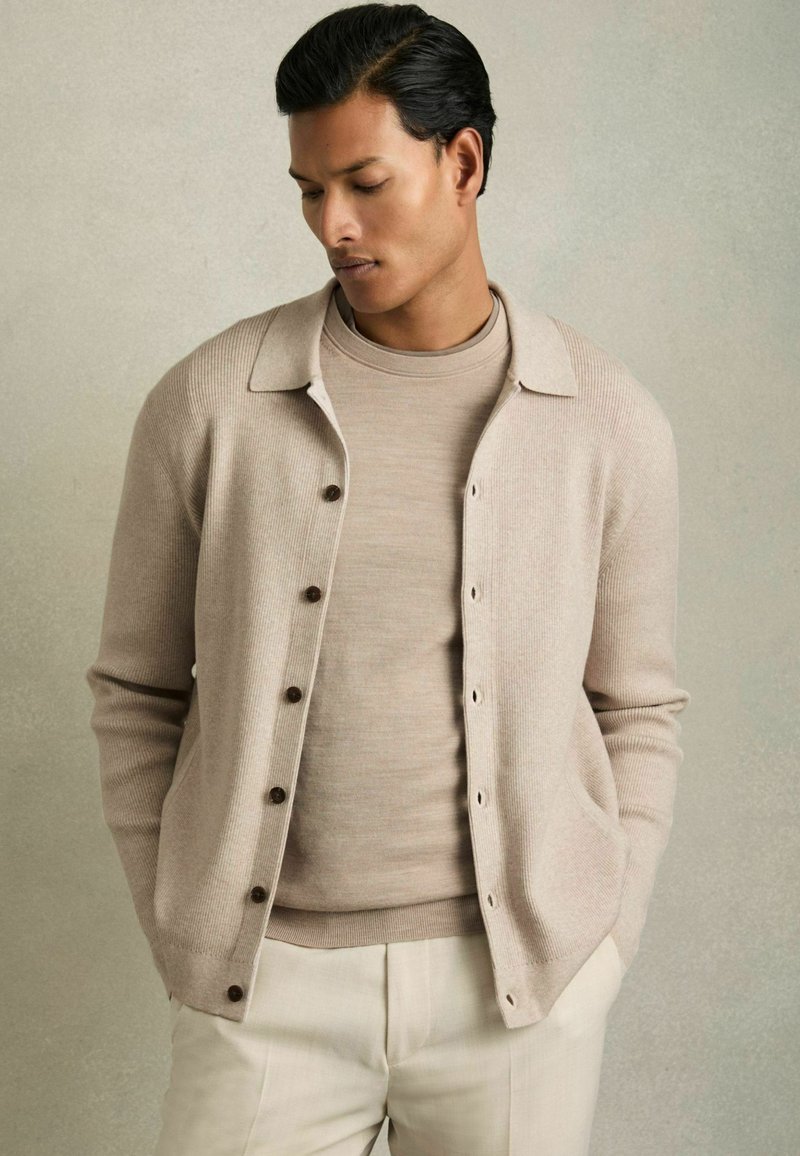 Beige cardigan med ribbat mönster, knappar och krage, lagerpålagd över en matchande polotröja. Ljust färgade byxor fulländar outfiten.