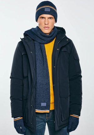 Jaunas vyras dėvi tamsiai mėlyną mezgimo kepurę, šaliką, pirštines, juodą žieminę striukę, geltoną megztinį ir mėlynus džinsus, stovintis prieš paprastą foną.