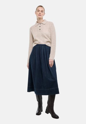 Femme portant un pull beige en maille torsadée avec des boutons, une jupe mi-longue plissée bleu foncé et des bottes noires montantes, se tenant devant un fond blanc.