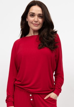 Rotes Langarmoberteil mit lässigem Schnitt, weichem Stoff und Bootsausschnitt. Kombiniert mit roten Hosen, die weiße Knopfakzente haben.